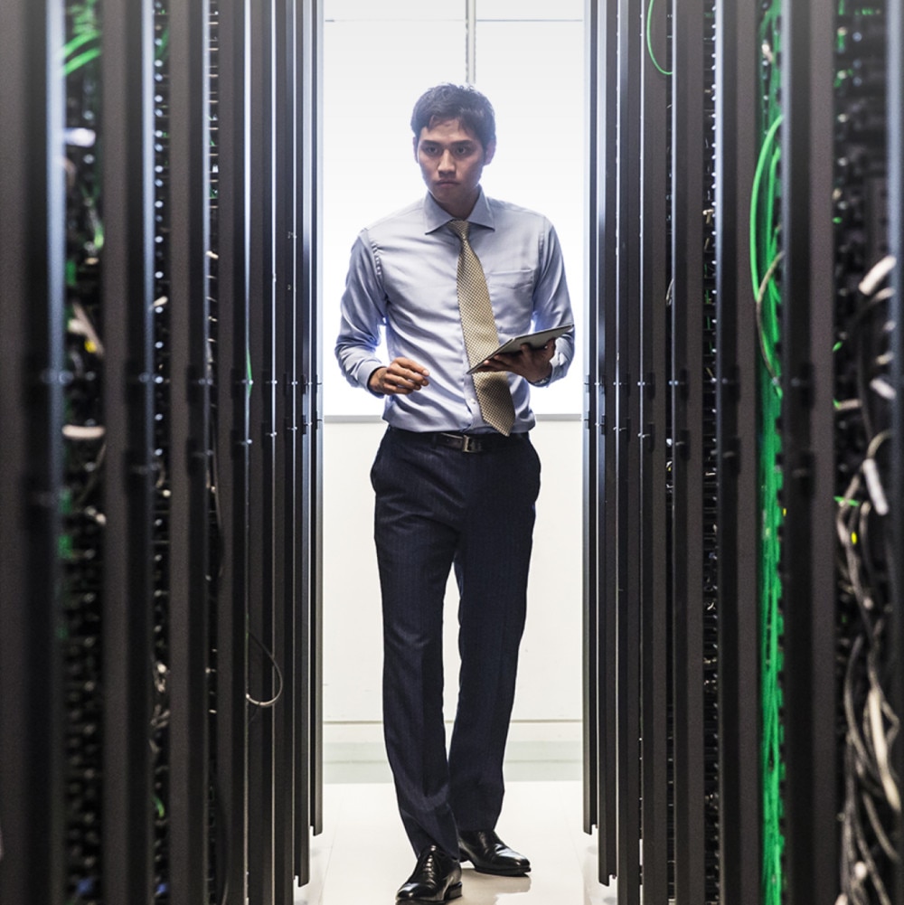 A person holding a notepad walks intently between aisles of server equipment.